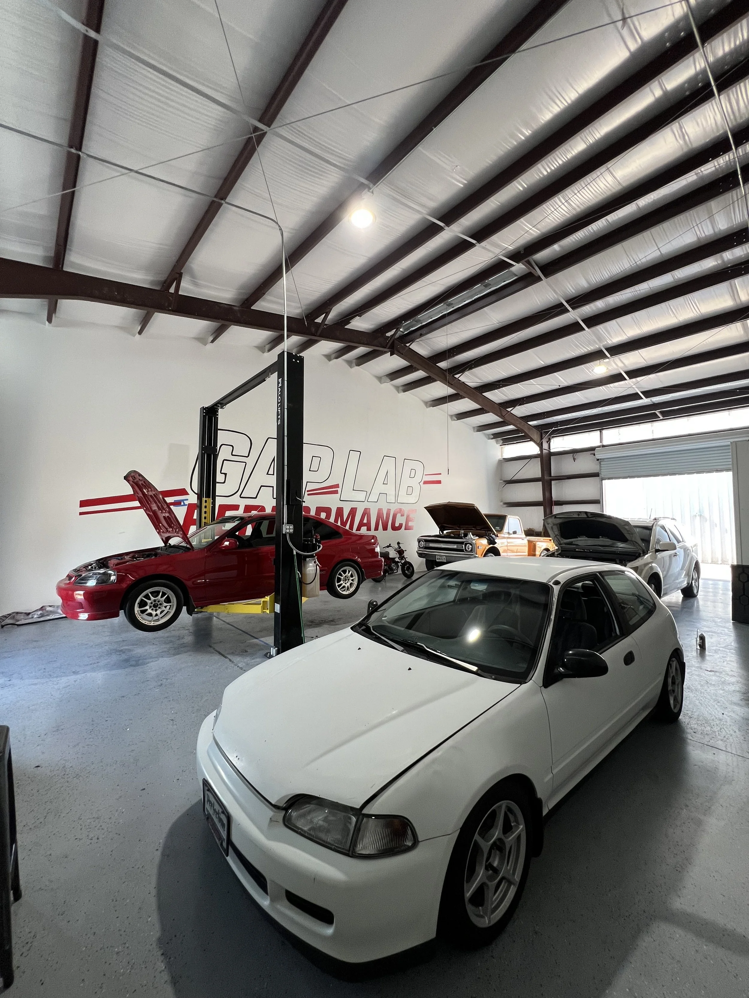 Inside a garage with several cars, including a white compact car in the foreground, a red car on a lift with an open hood, and two vehicles with open hoods in the background, along with a sign reading "GAP LAB PERFORMANCE."