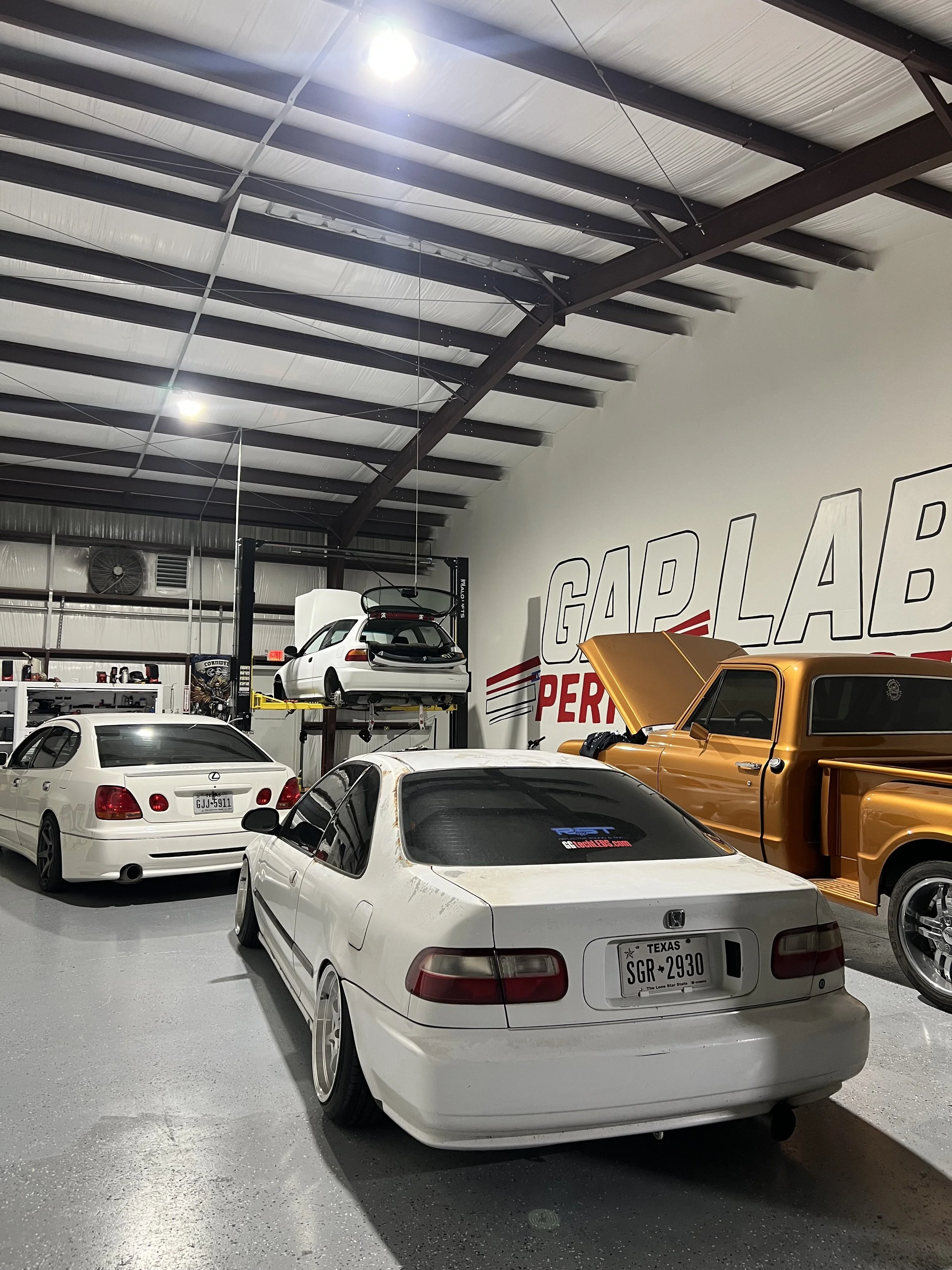 An indoor garage with several cars, including a white Honda with a Texas license plate, a white Lexus, a gold truck with its hood open, and a white car lifted on a hydraulic lift. The garage has a high ceiling with metal beams and a sign on the wall that reads 'GAP LAB PERFORMANCE'.