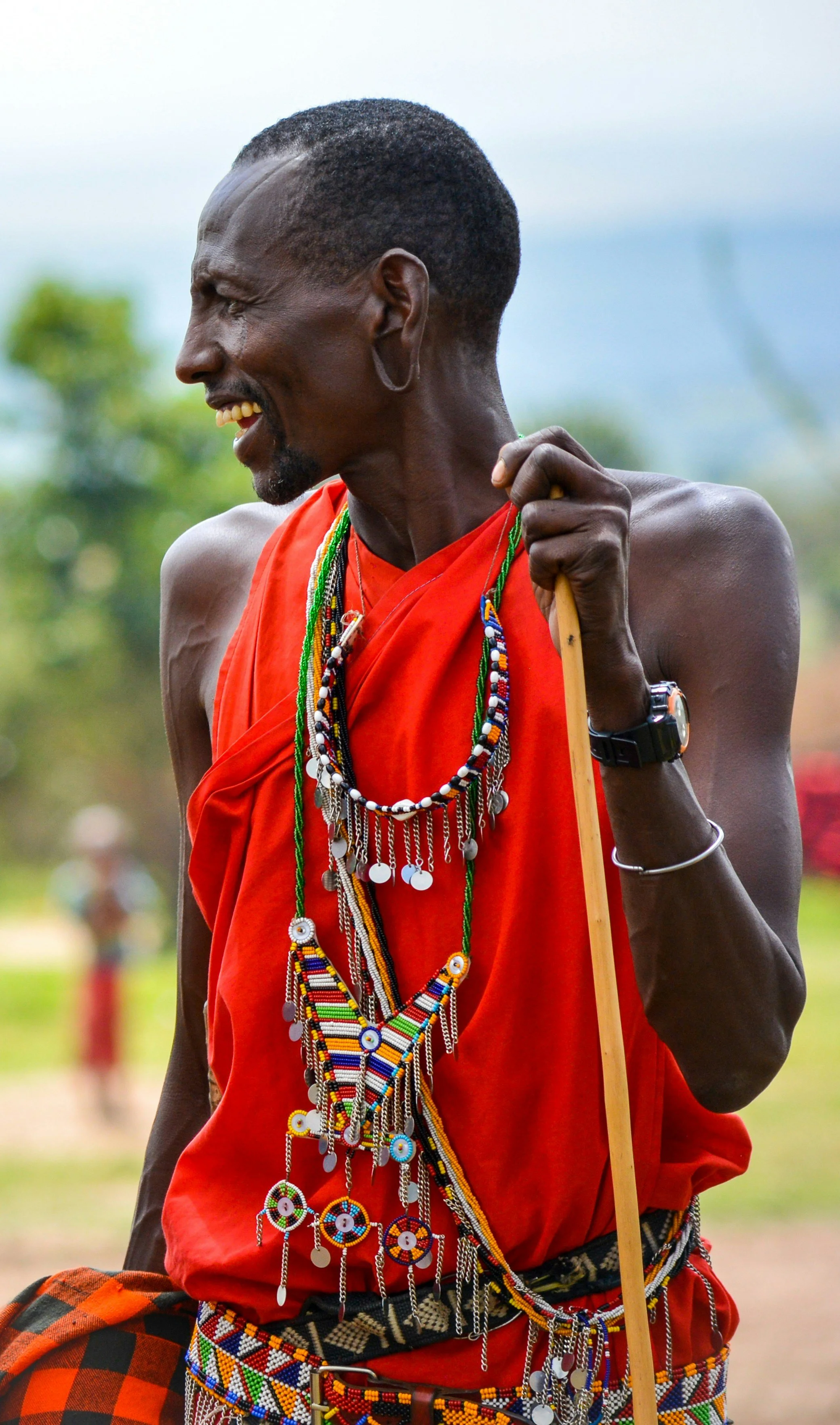 A man dressed in traditional Maasai attire and jewelry, holding a wooden staff, smiling outdoors.