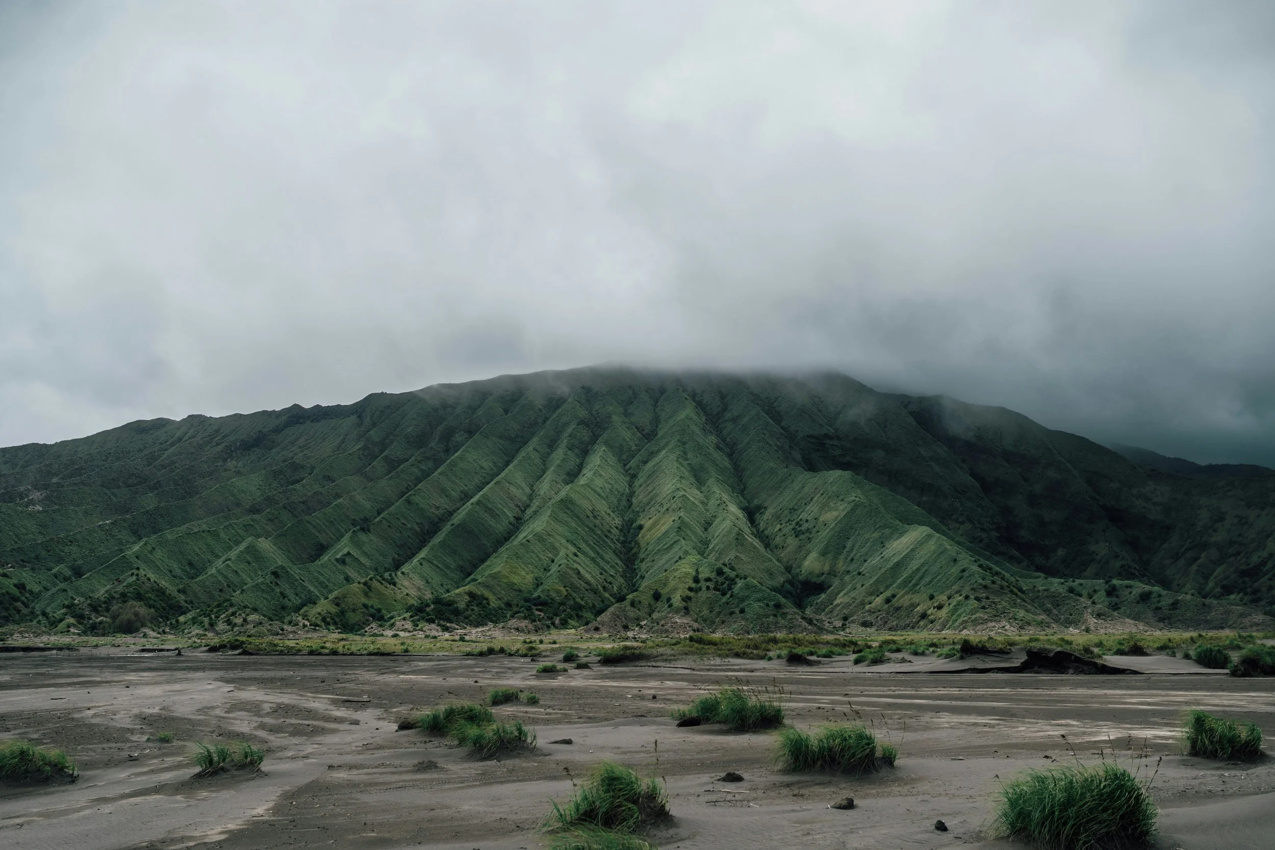 A green mountain with a cloud-covered peak and patches of green vegetation in the foreground, along a dry, sandy area.