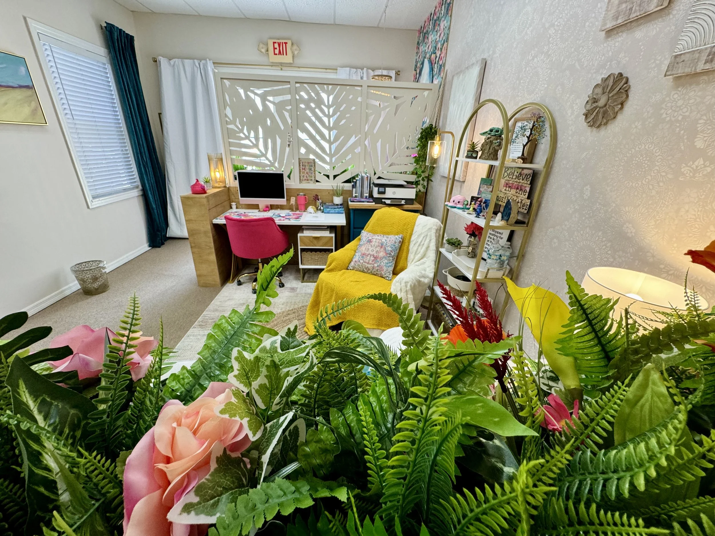 A cozy therapy office with a floral arrangement in the foreground, a desk with a monitor, pink chair, books, and decor, and a shelving unit displaying various decorative items.