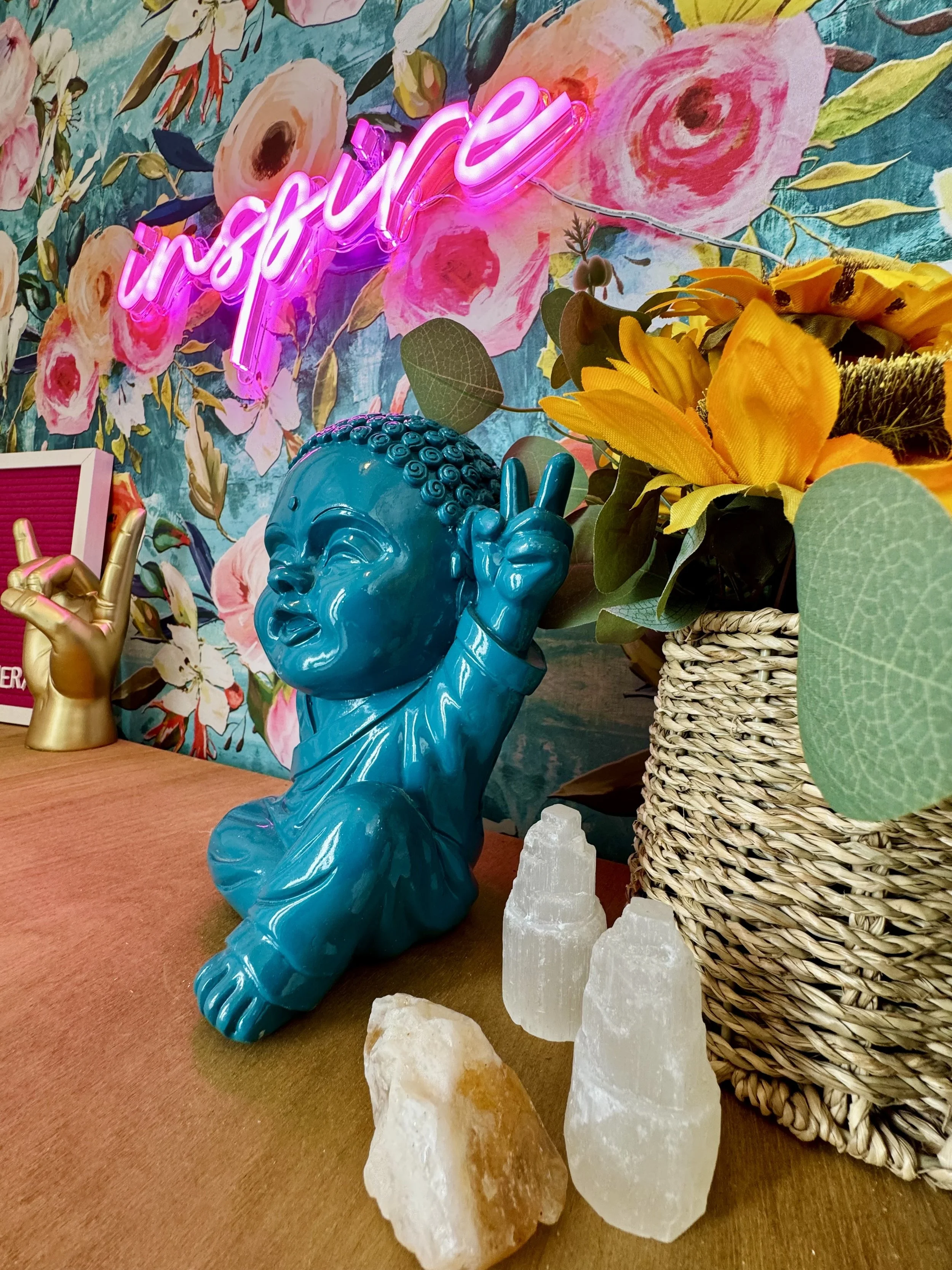 Decorative display with a bright blue Buddha figurine making a peace sign, surrounded by a sunflower bouquet in a woven basket, Himalayan salt crystals, a gold hand sculpture, and a pink neon sign