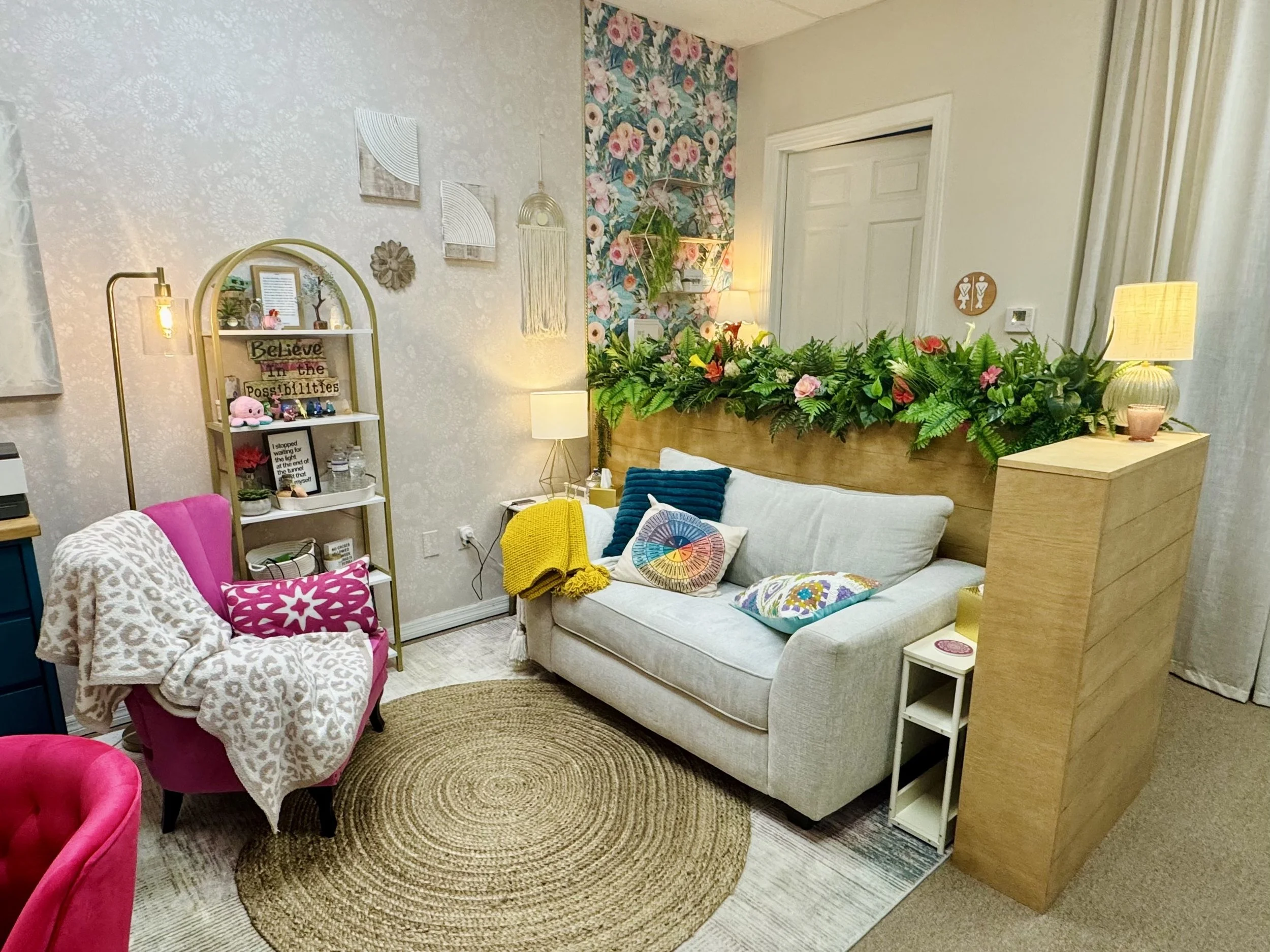 Cozy therapy room with a beige sofa, colorful pillows, a yellow blanket, and a floral photo wall. A pink armchair with a leopard print blanket, a circular rug, and a tall white shelf with decorations.