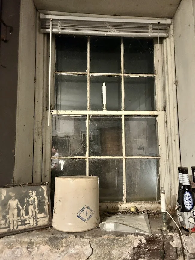 Old, dirty window with a closed blind, surrounded by dust and items on a worn surface, including a photograph, a white container, and some bottles.