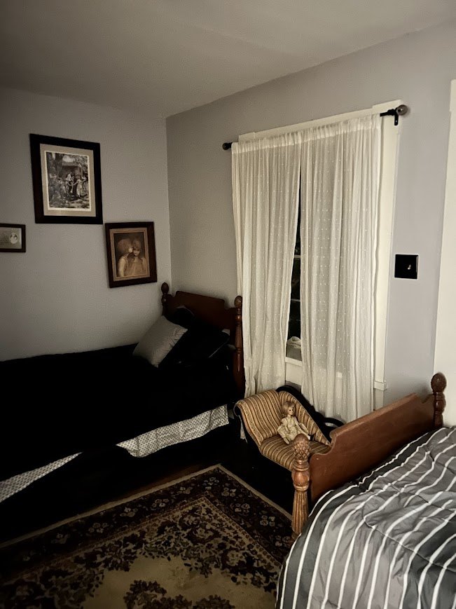 A bedroom with two wooden beds and a small striped armchair. The bed on the right has a striped blanket, and the bed on the left has dark linens with pillows. There are framed pictures on the wall and a window with white curtains.