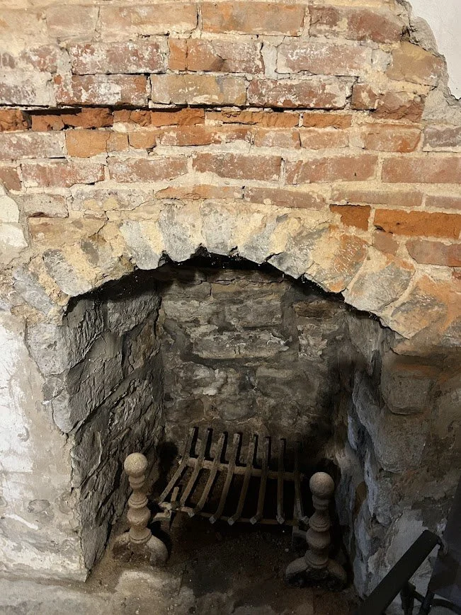Close-up of an old brick fireplace with a small wooden grate inside, set in a stone and brick wall.