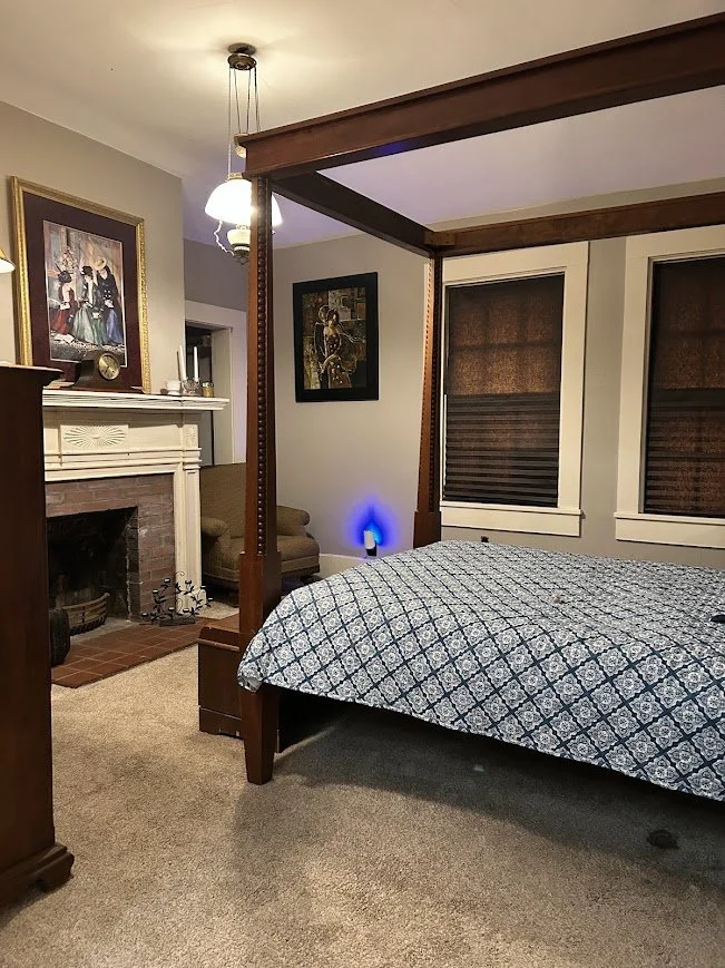 A cozy bedroom with a four-poster bed, fireplace with artwork above, armchair, and window blinds, warmly lit with ceiling light and blue accent lighting.