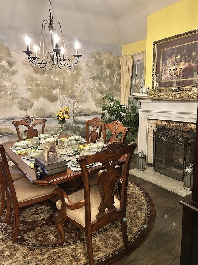 A dining room with a wooden table set for six, surrounded by carved wooden chairs with upholstered seats. The table has a flower vase with yellow flowers, place settings, and glasses. There is a large rug underneath the table. A chandelier hangs from the ceiling, and a fireplace with a framed painting above it is on the right side. A large plant and a window with curtains are in the background.