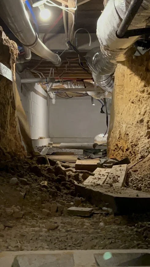 View of an underground utility or construction site with pipes, gravel, and dirt, and an unfinished ceiling with visible wood and electrical wiring.