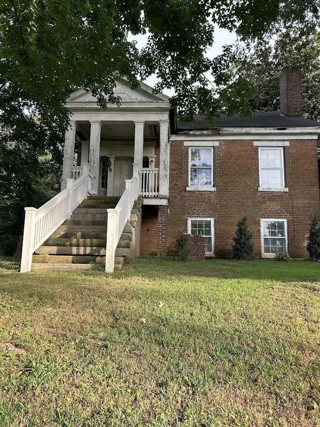 A brick house with a front porch accessed by a staircase, surrounded by a grassy yard with some small shrubs and trees