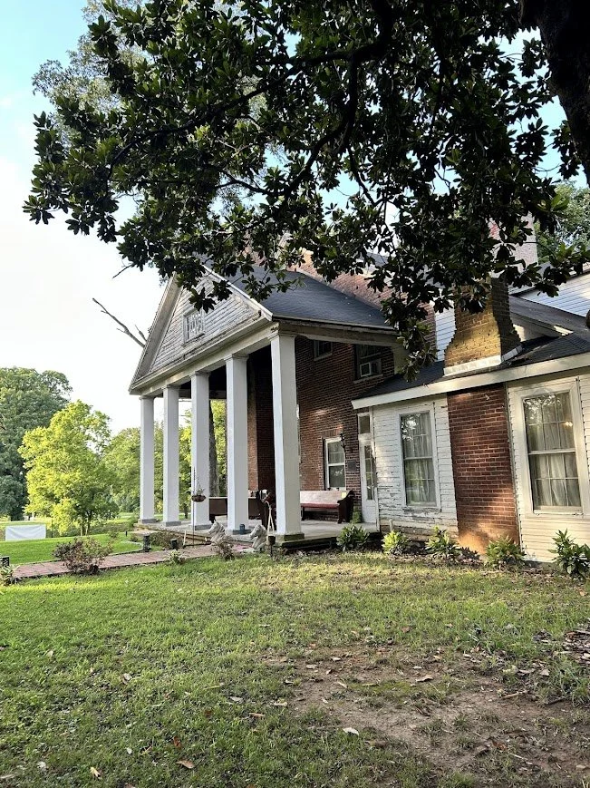 A large house with white columns and a brick exterior, surrounded by greenery and trees.