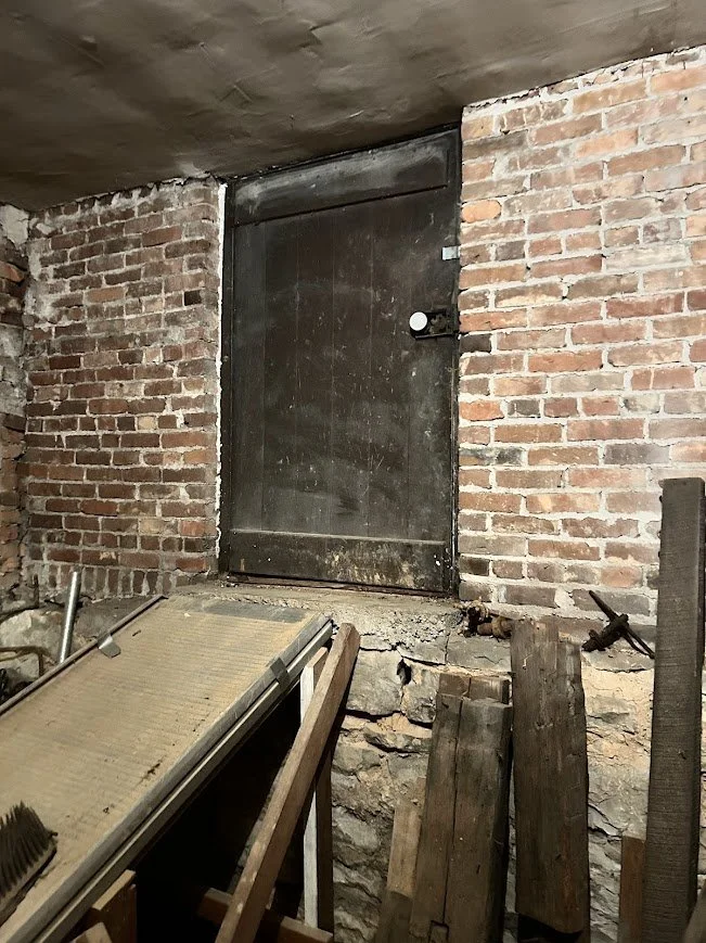 A black metal door with a white doorknob, set in an exposed brick wall, in a basement or cellar with construction materials and tools nearby.