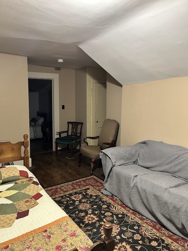 View of a cozy living room with a bed partially visible on the left, a patterned rug, a gray-covered sofa, and two wooden chairs near a doorway leading to a bedroom.