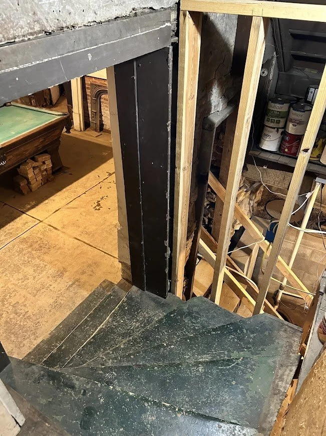 View of a basement stairwell under construction with wooden framing and concrete staircase, showing construction materials and household items in the background.