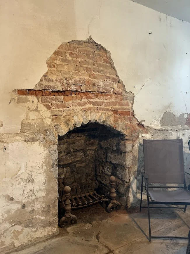 A partially exposed brick fireplace with old stone and brickwork inside and a damaged plaster wall surrounding it, located next to a patio chair.