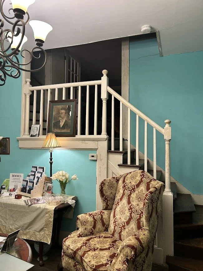 Living room with light turquoise walls, a vintage patterned armchair, a table with books and flowers, a floor lamp, and framed portraits on the wall. A staircase leads to a second floor with a railing.