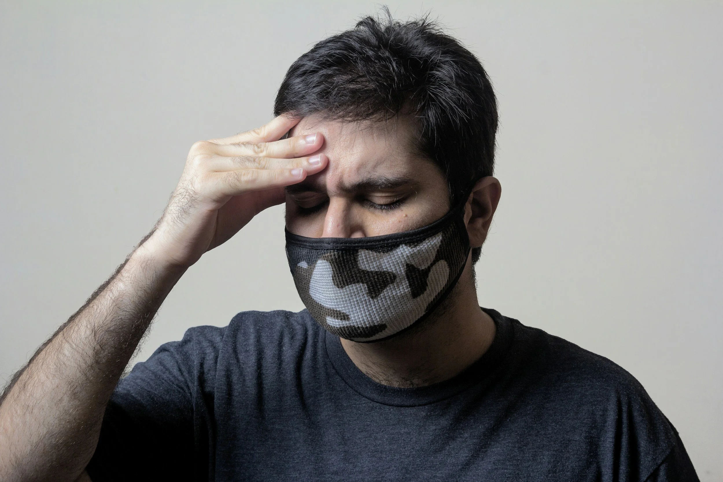 A young man with dark hair wearing a camouflage face mask, holding his forehead with one hand, appears to be experiencing a headache or discomfort, against a plain light background.