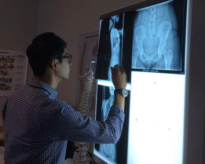 A male doctor examining spinal X-ray films on a lightbox in a medical office.