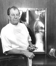 A man in a white coat holding an X-ray film, standing in front of an X-ray display with images of a skull and a foot.