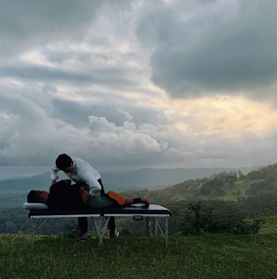 A person receiving a massage outdoors on a portable massage table with a scenic mountainous landscape and cloudy sky in the background.