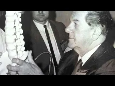 A man in a suit holding a model of a human spine, possibly at a medical or dental event, with another man observing.