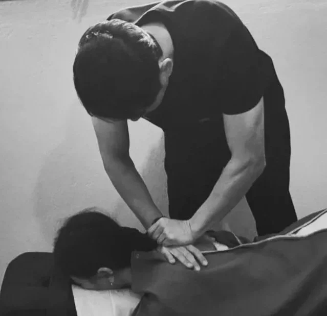A person leaning over another person who is lying face down on a bed or table, with one hand on their shoulder and the other on their head.