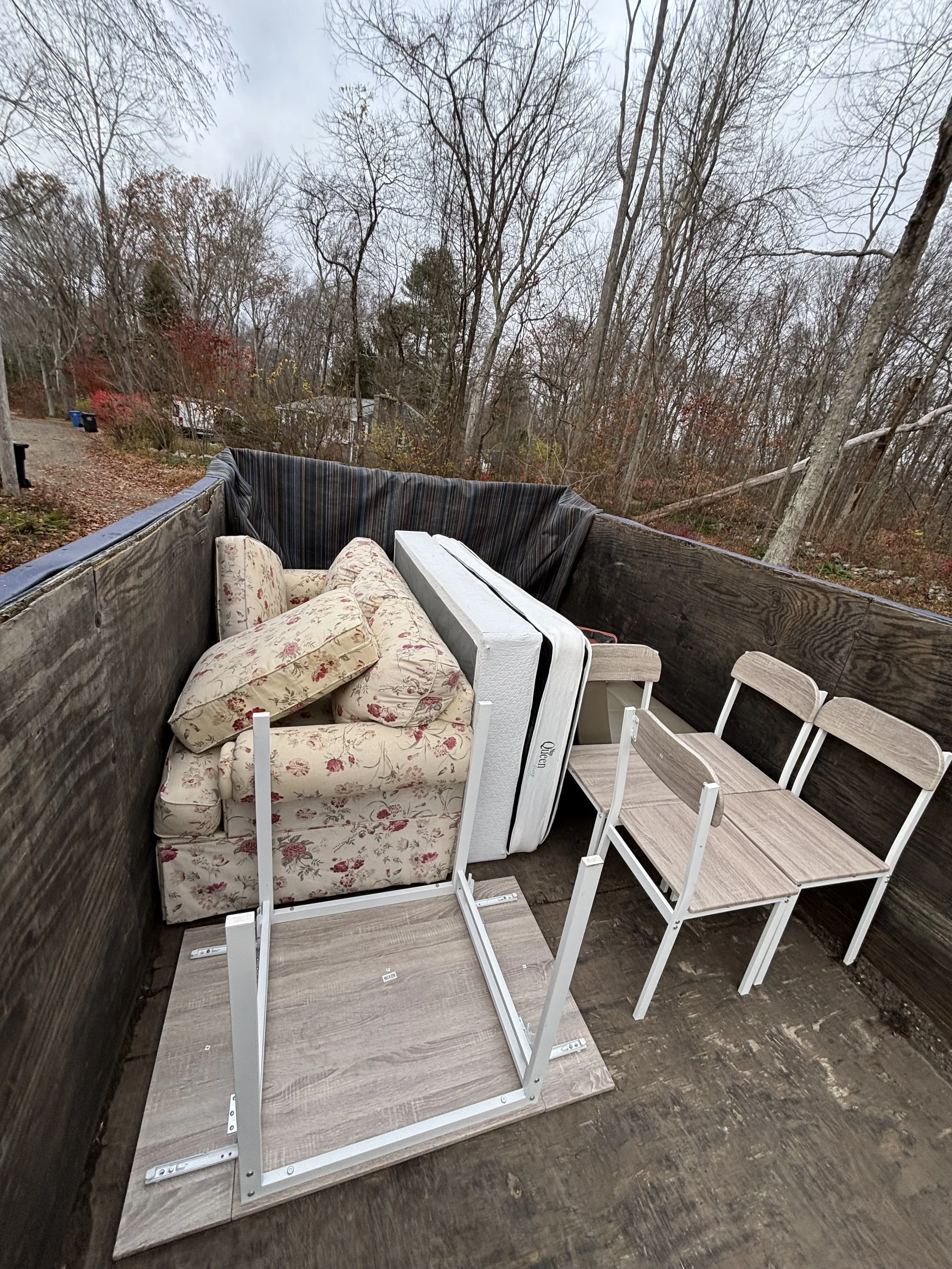 The image shows a porch or deck area with a wood floor and a wooden railing. There are several pieces of furniture stacked together, including a floral upholstered armchair, an upholstered loveseat, a small table, and several small wooden chairs. In 