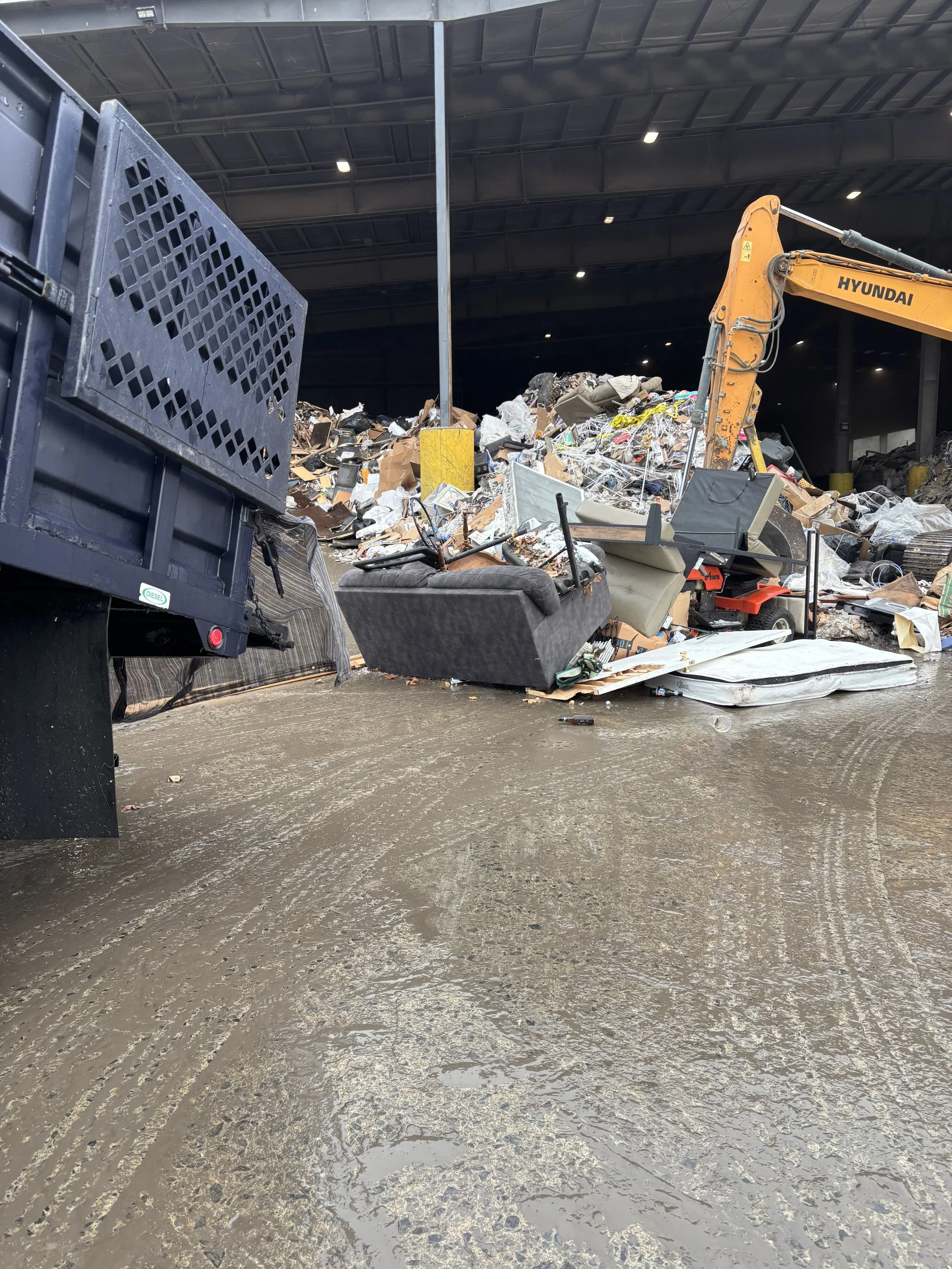 Dump truck unloading a large pile of mixed debris, including furniture and household items, inside a warehouse or recycling center with a Hyundai excavator in the background.