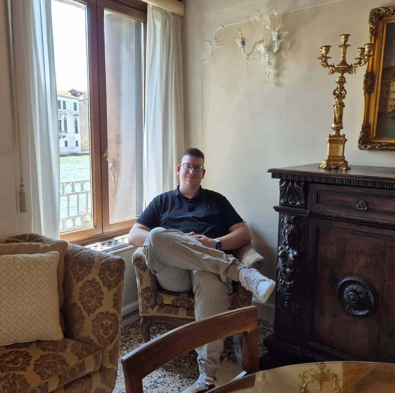 Ein junger Mann sitzt entspannt auf einem gepolsterten Sessel vor einem großen Fenster mit weißen Vorhängen in einem eleganten, klassisch eingerichteten Raum. Der Raum ist dekorativ mit antiken Möbeln und einem goldverzierten Bilderrahmen sowie einem Kerzenständer aus Gold ausgestattet.