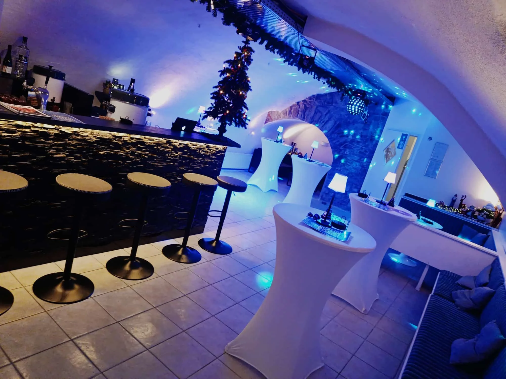 Ein festlich dekorierte Lounge mit einer Bar, einem kleinen Weihnachtsbaum, Stehtischen mit weißen Hussen, LED-Beleuchtung und blauer Lichtstimmung, geeignet für eine Weihnachtsfeier.