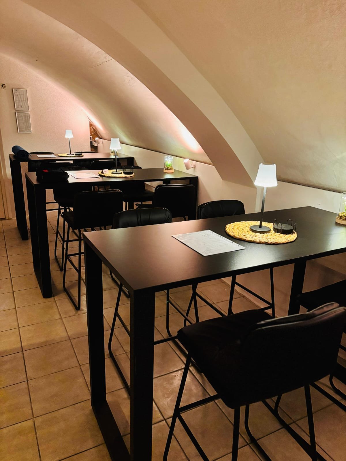 Ein Raum mit schräg verlaufender Decke, in dem mehrere schwarze Tische mit schwarzen Stühlen stehen. Auf den Tischen befinden sich kleine Lampen, Dekorationen und Menükarten.