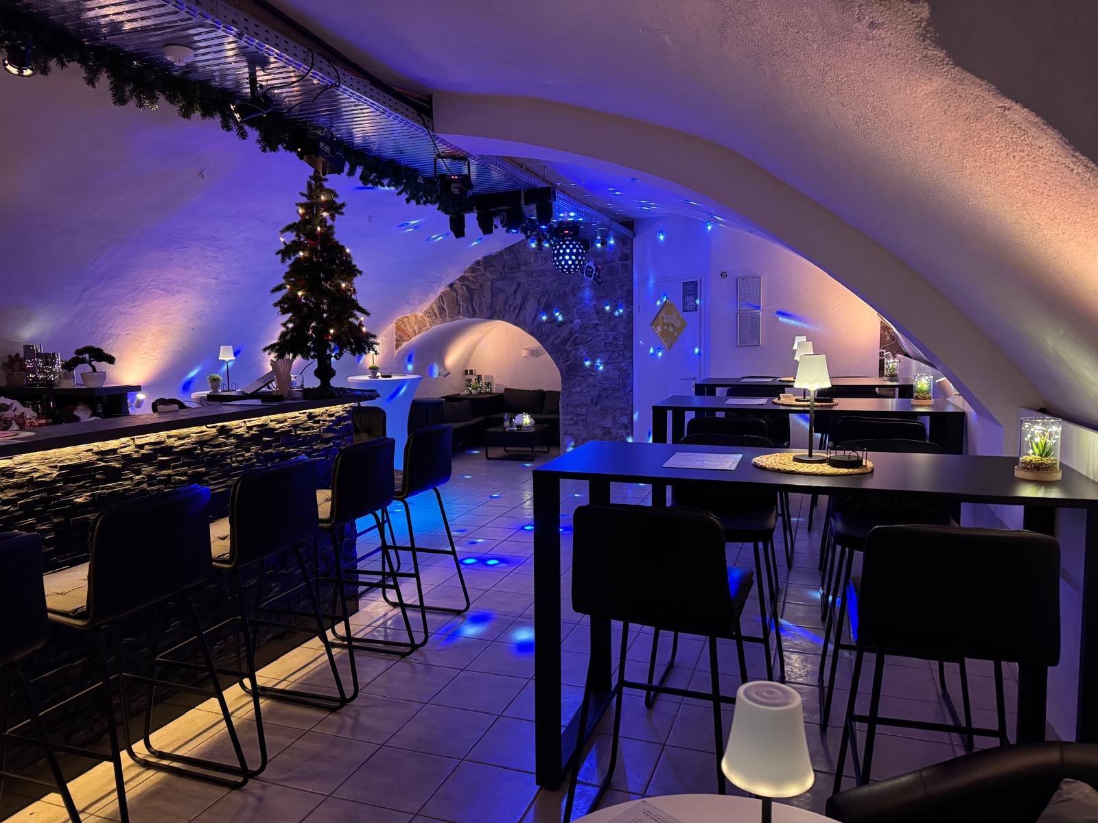 Gemütliche, modern gestaltete Bar oder Lounge mit Weihnachtsdekoration und blauen Lichtakzenten, inklusive Weihnachtsbaum, kleinen Tischlampen und Dekorationsgegenständen auf Tischen.