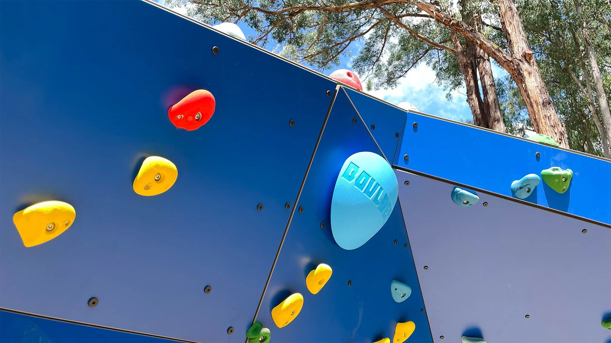 Colorful outdoor climbing wall with red, yellow, blue, and green handholds, set against a blue sky with trees in the background.