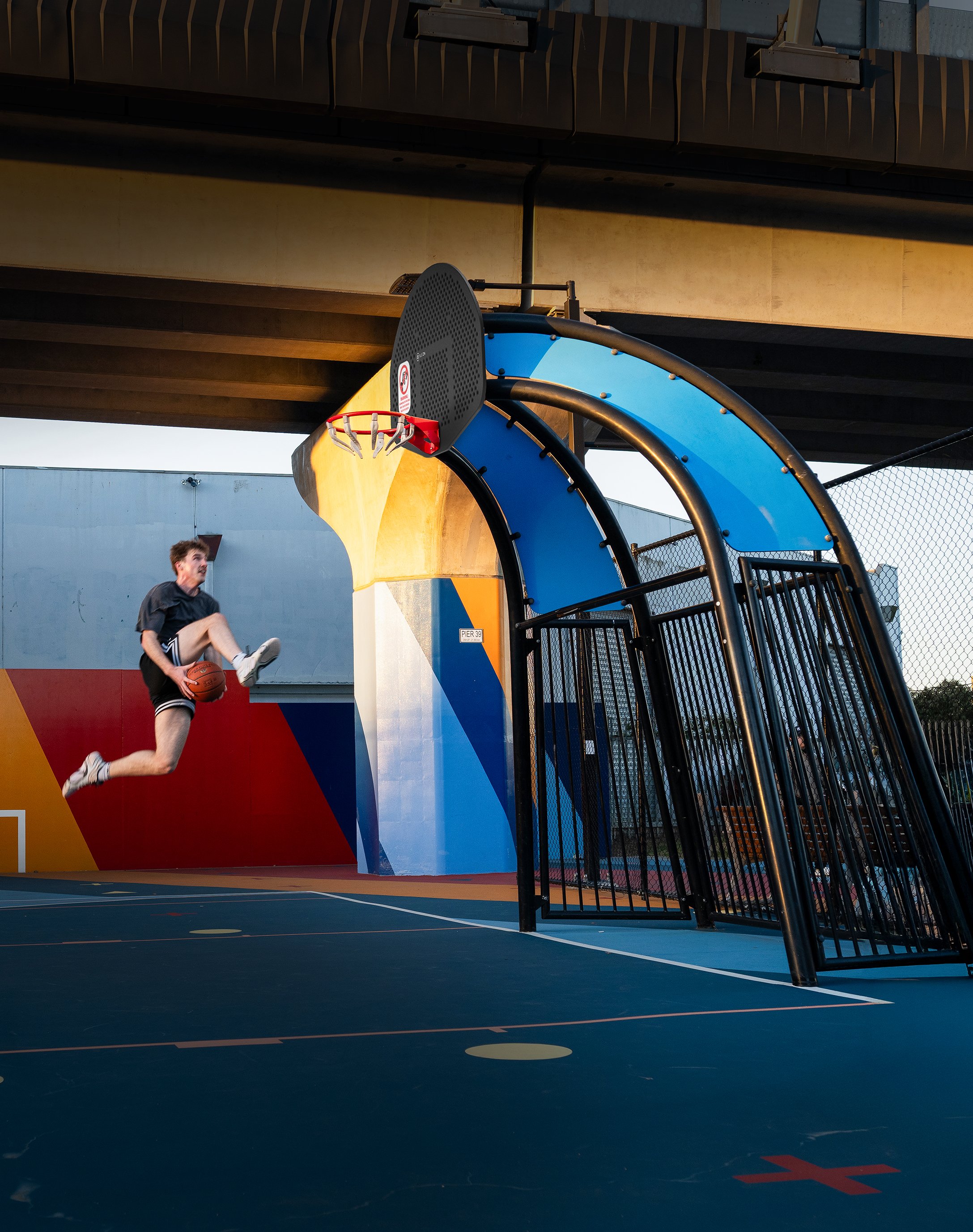 A person dunking a basketball on an outdoor court under an overpass during sunset.
