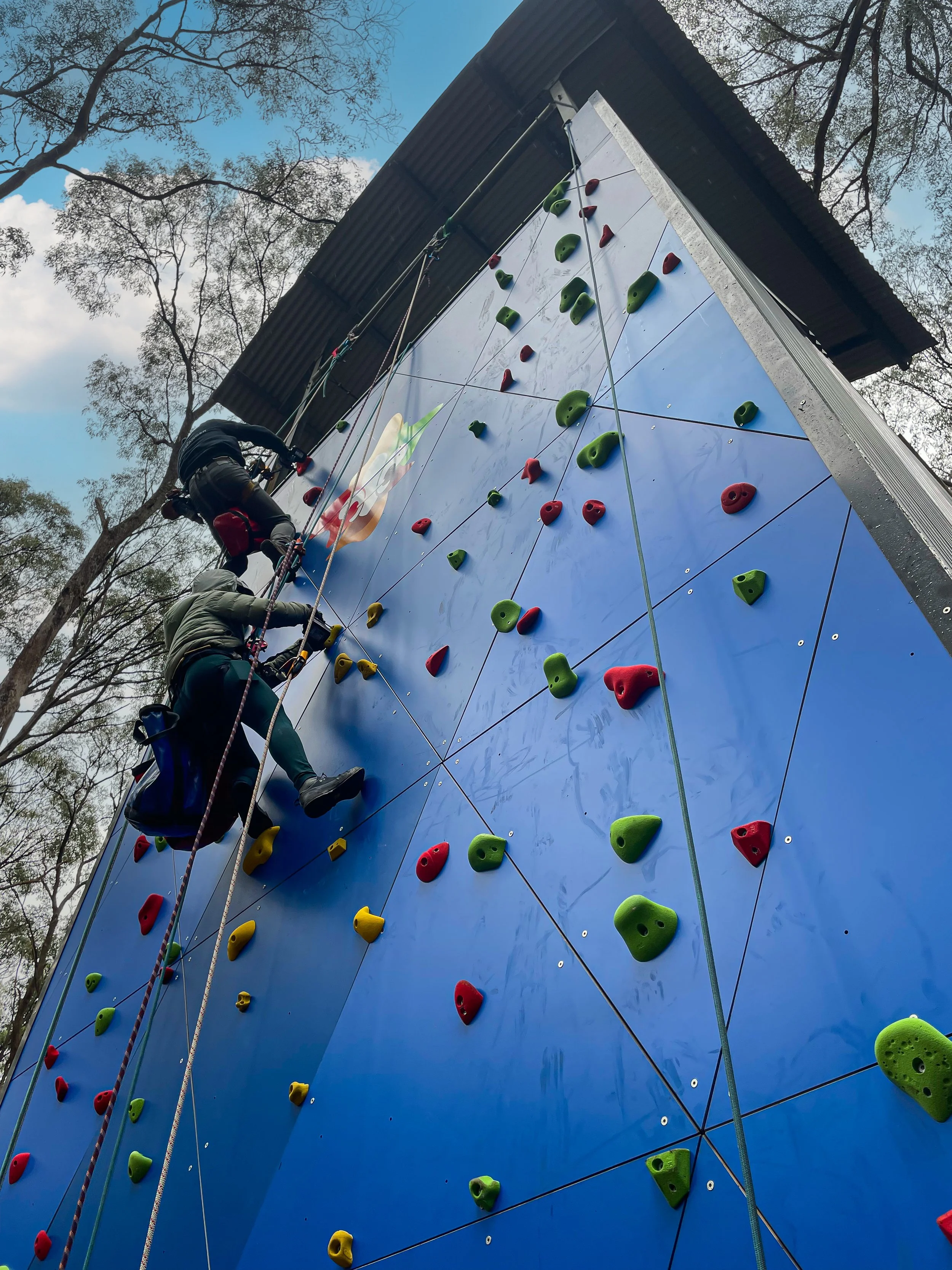 Two people climbing an outdoor bouldering rock climbing wall with colorful holds, in a natural setting with trees and blue sky.
