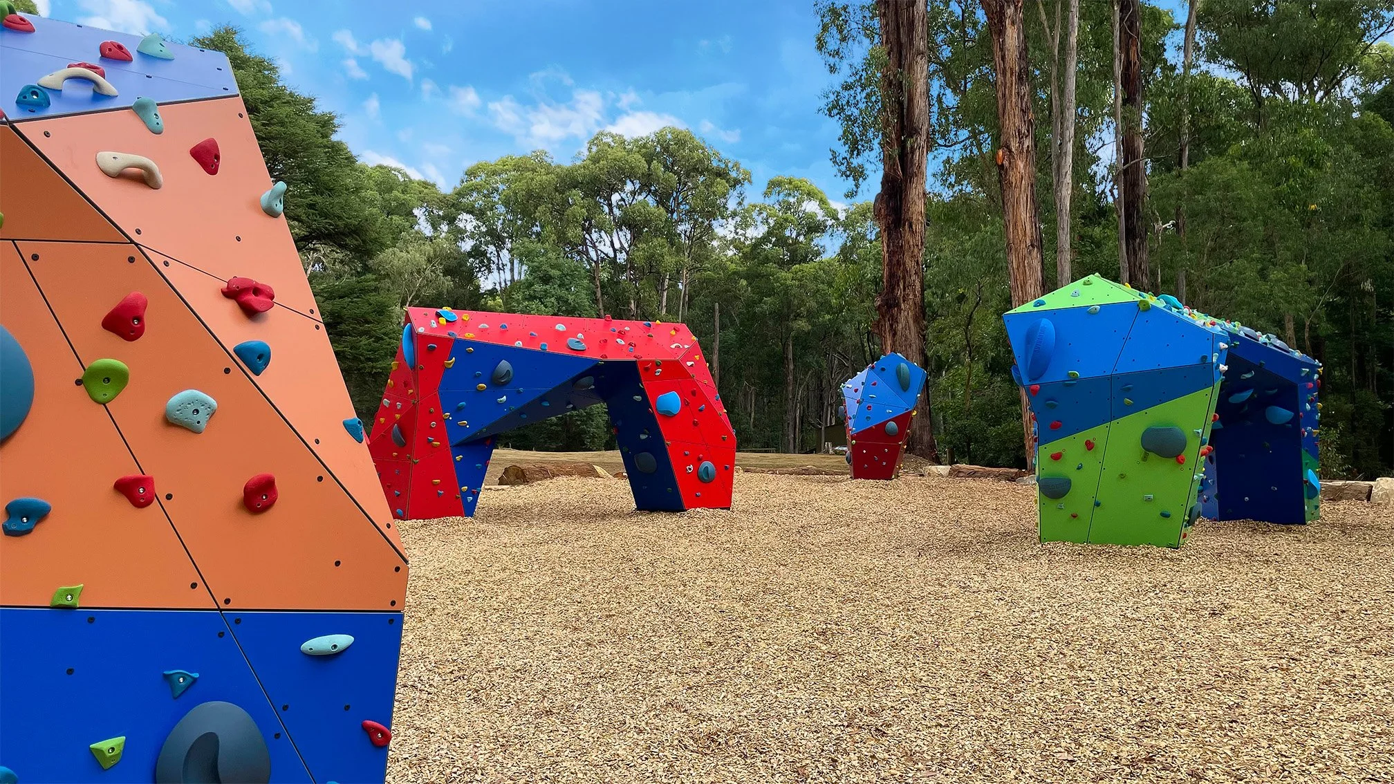 Colorful outdoor climbing structures and boulders in a wooded park setting.