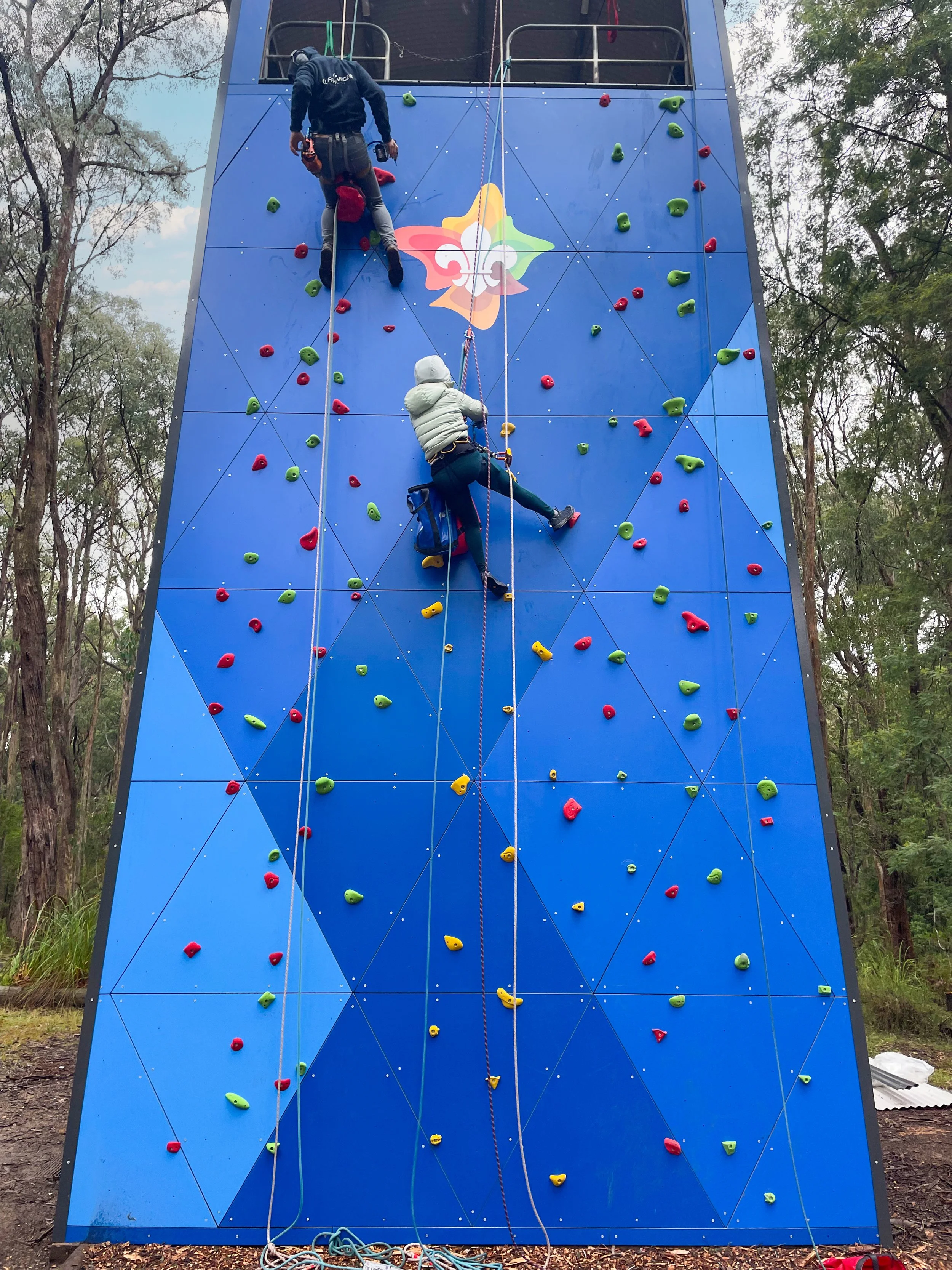 Two people are climbing a blue outdoor rock climbing wall with colorful holds, in a wooded area. One climber is higher and being belayed by a person below.