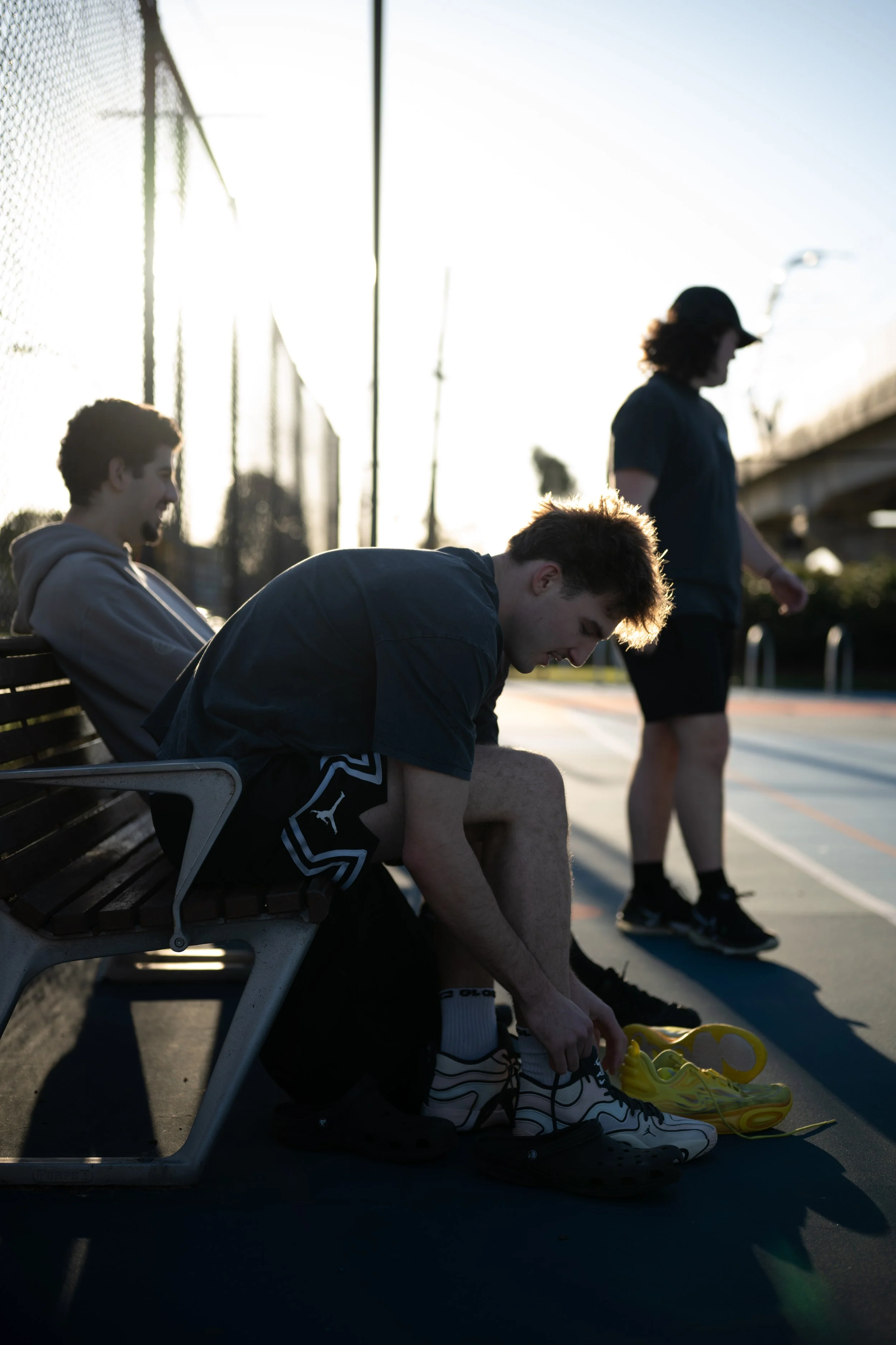 Three young men at an outdoor basketball court, with two sitting on a bench and one standing, as the sun sets behind them.