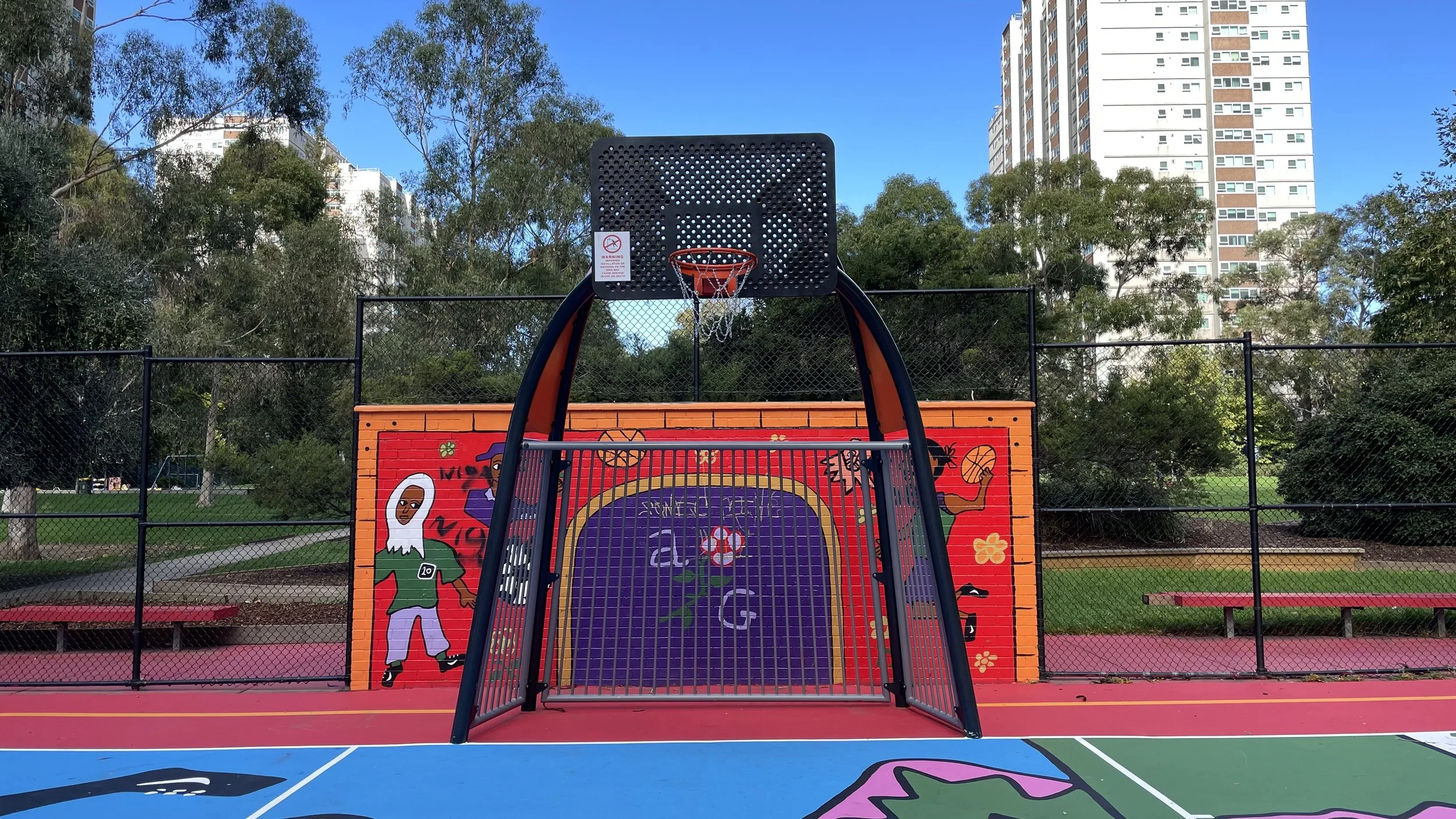 Fusion basketball tower on a colorful playground with painted walls and trees, high-rise buildings in the background.