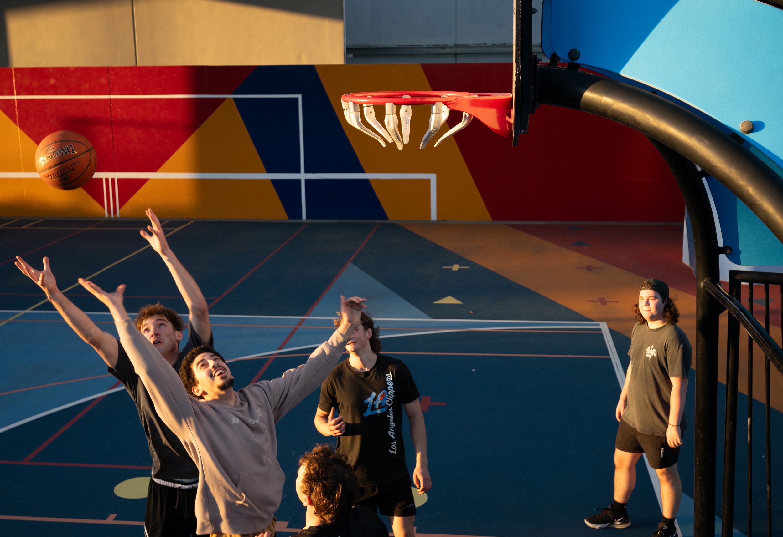 Four teenagers playing basketball on an outdoor court during sunset, with a basketball in mid-air near the hoop. Includes a Fusion Tower and Infinet Basketball ring