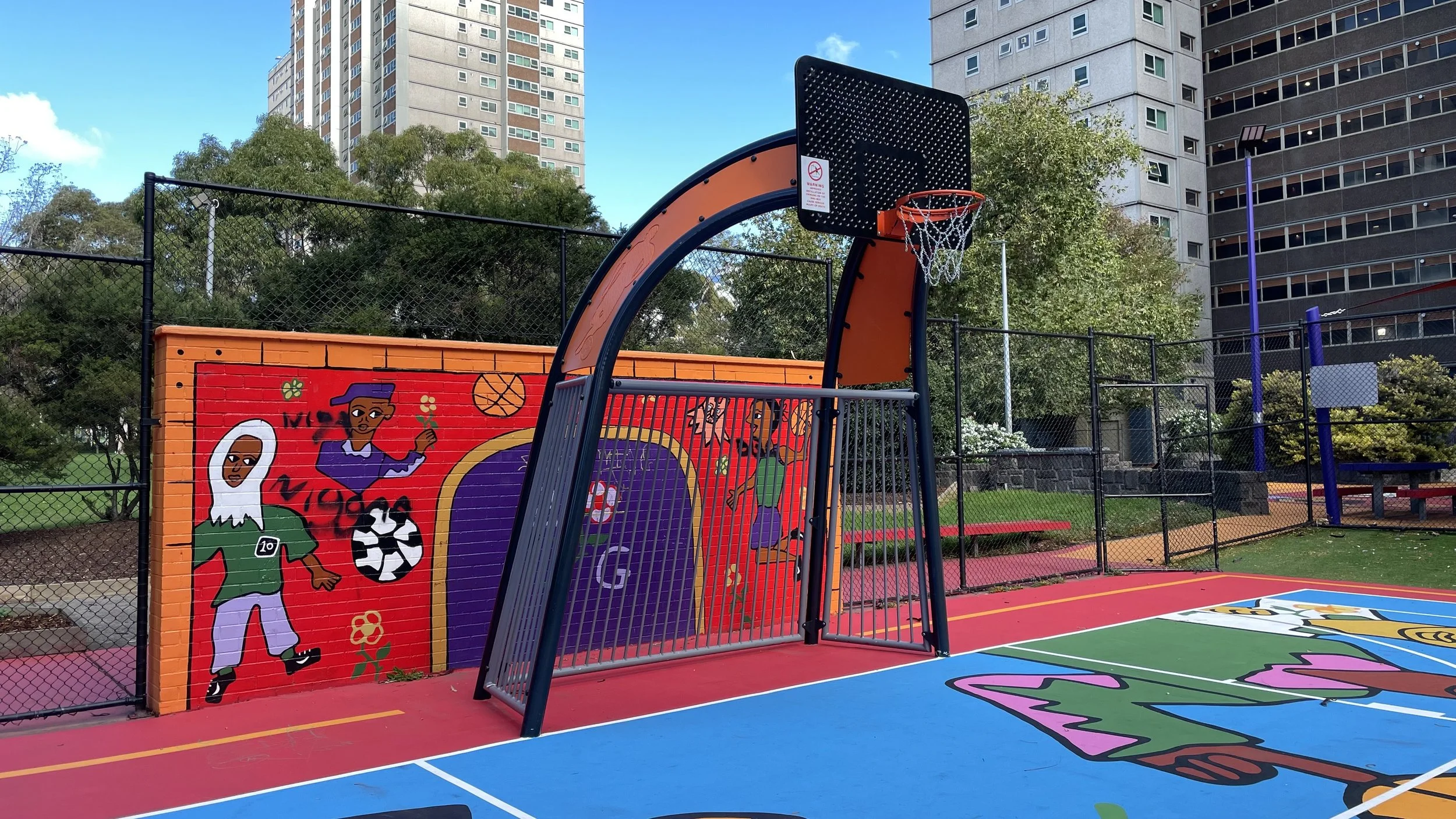 Fusion basketball tower on a colorful playground with painted walls and trees, high-rise buildings in the background.