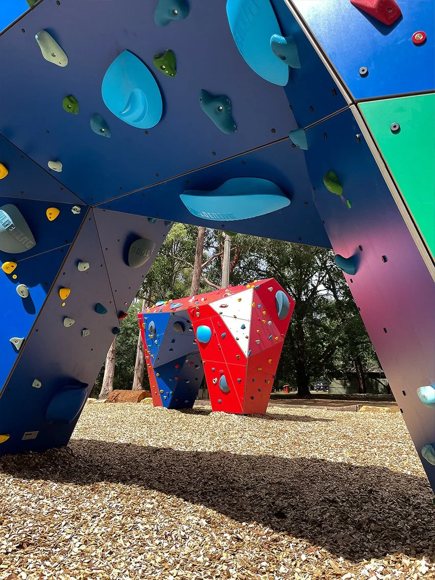 Colorful outdoor climbing structures with various holds, set on a ground covered in wood chips, in a park with trees.