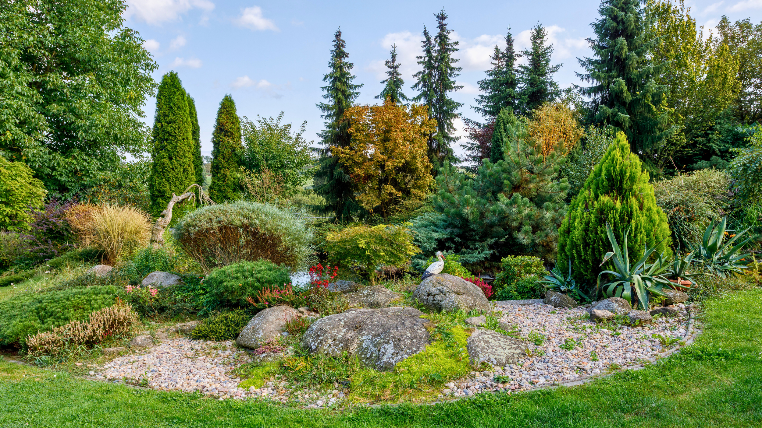 Colorful lush garden with various trees, shrubs, and rocks, featuring a white bird perched on a large rock in the center, under a partly cloudy sky.