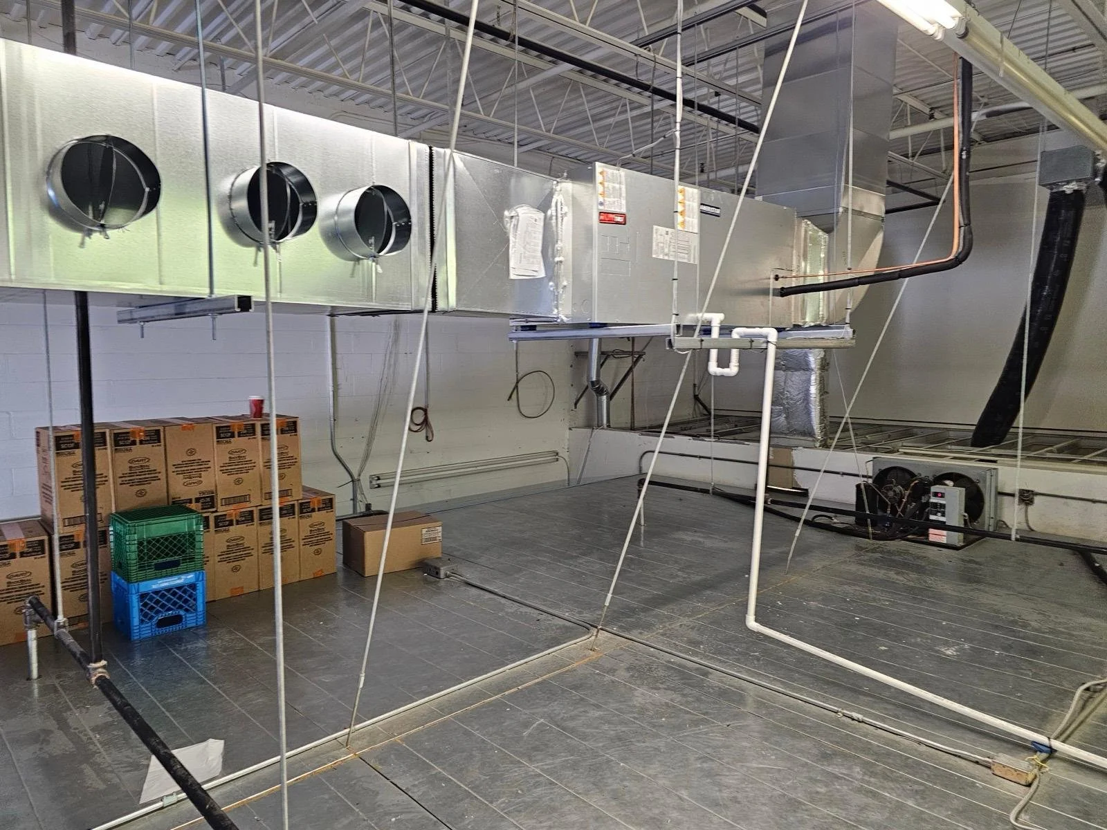 Empty industrial kitchen or food service area with boxed supplies, empty flooring, and large stainless steel HVAC units or ventilation system overhead.
