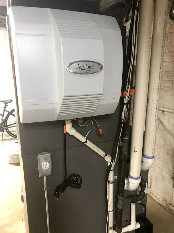 A white Aprilaire humidifier mounted on a black wall with electrical wires and white pipes nearby.