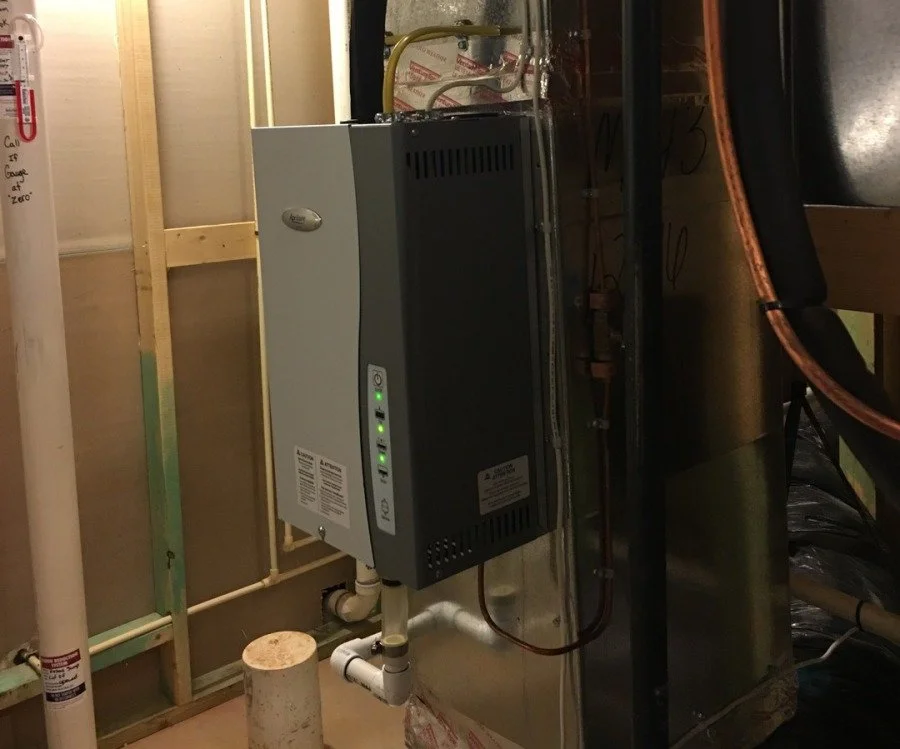 A small electrical or HVAC unit mounted on a wall in a basement or utility room, with pipes, wiring, and unfinished wall studs visible around it.