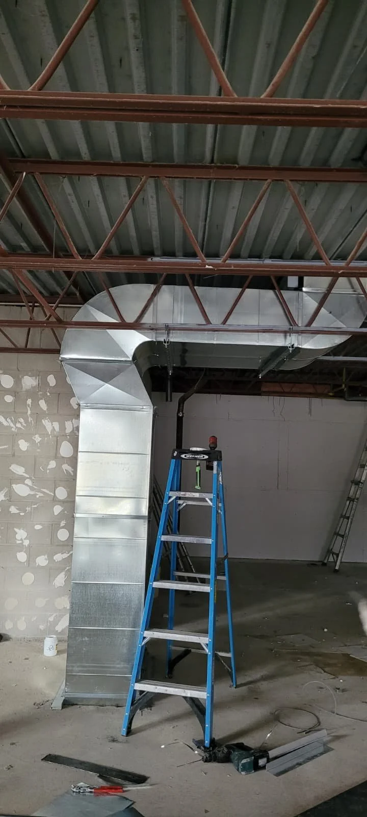 Interior construction site with metal ductwork, a blue step ladder, and construction tools on the floor.
