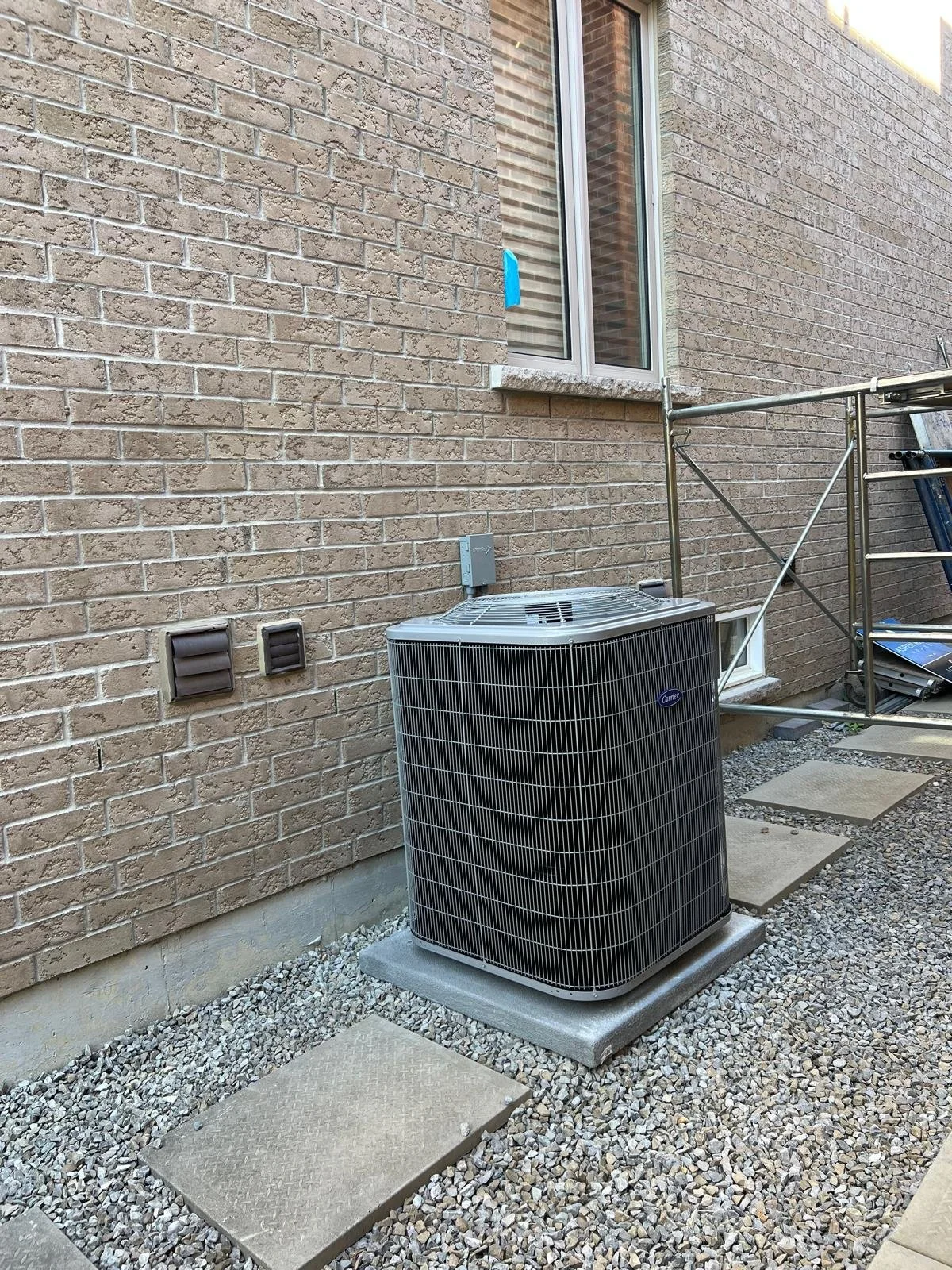 The image shows the exterior of a house with a brick wall, a window, and an outdoor air conditioning unit. The ground is covered with gravel and concrete stepping stones. There is scaffolding next to the house.