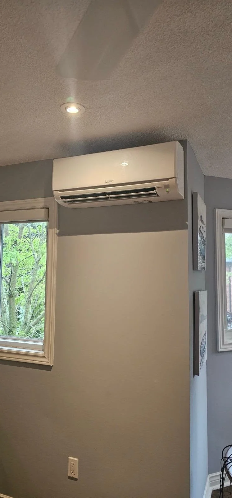 A wall-mounted Mitsubishi air conditioner in a room with a window showing green trees outside.