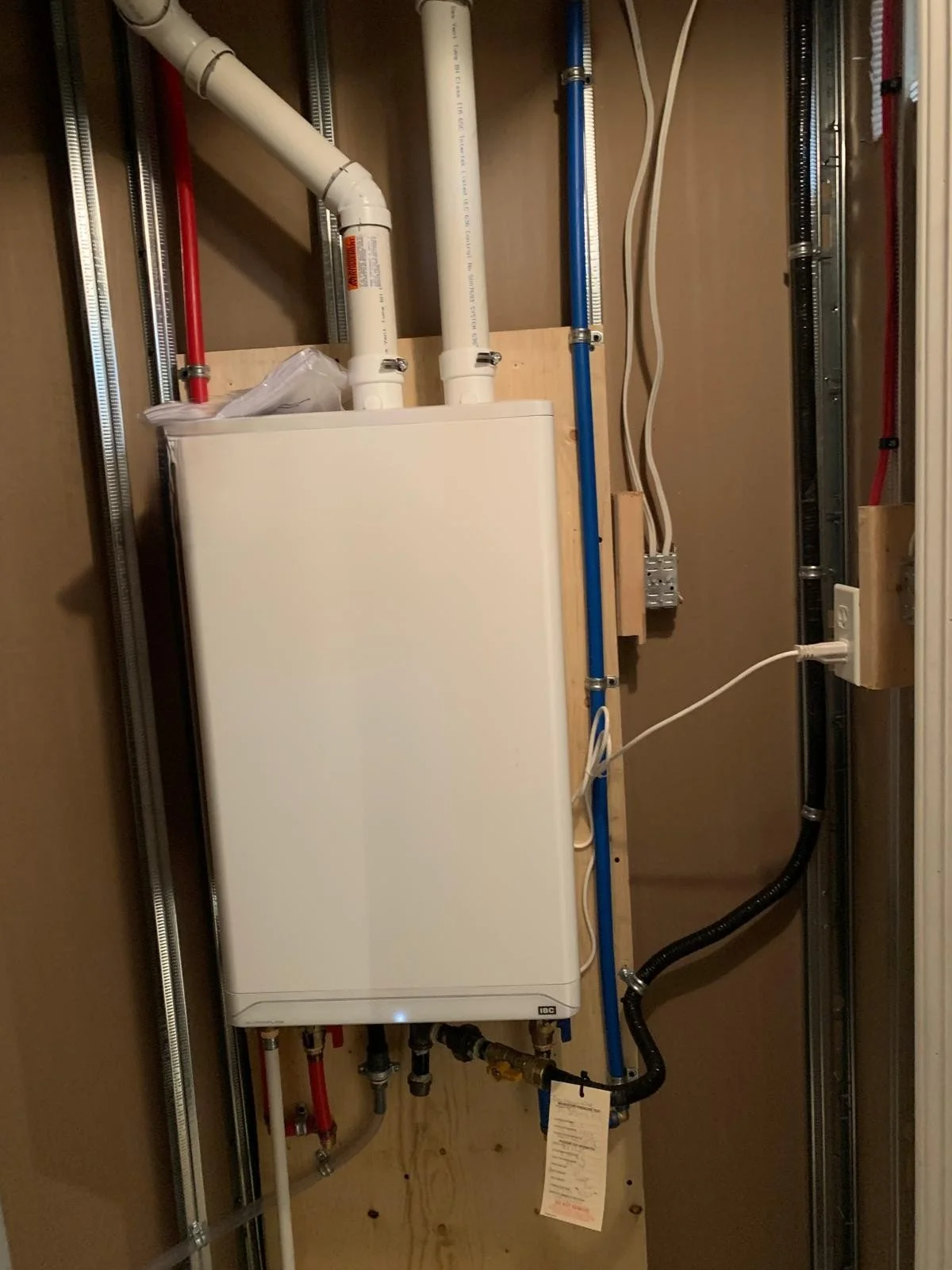 A water heater installed in a utility closet with various pipes, including red, white, and blue, connected to it. The setup includes electrical wiring and a black flexible pipe, with the heater mounted on a wooden panel.